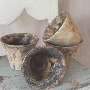 May include: Three weathered, conical pots with a rough, textured surface in shades of brown, beige, and charcoal. The pots feature a distressed, aged appearance with visible dark staining and uneven rims, arranged in a cluster with one tilted forward.