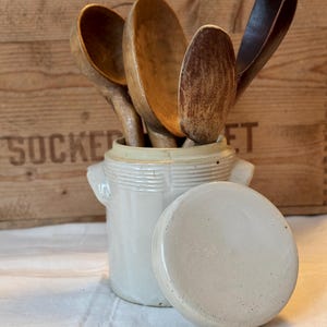 Op de afbeelding: Een crèmekleurige keramische pot met deksel, met houten lepels. De lepels variëren in kleur van licht- tot donkerbruin. De pot heeft twee kleine handvatten en horizontale ribbels. De achtergrond is een houten krat.