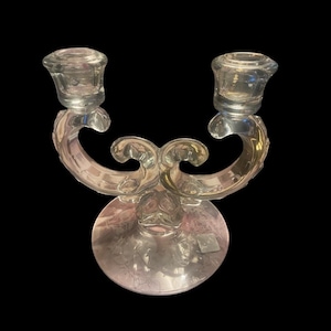 May include: Clear glass candelabra with two candle holders. The candelabra has a decorative, curved design with a round base. The candle holders are small and cylindrical. The overall design is ornate and elegant.