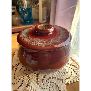 May include: A round, lidded ceramic jar in a deep red hue. The lid features a small, circular handle and a speckled, greenish-blue pattern. The jar sits on a white, lace doily.