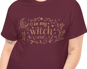 May include: A maroon t-shirt with a gold graphic that reads "in my witch era" with a crescent moon, stars, and floral accents. The shirt is worn by a person in blue jeans.
