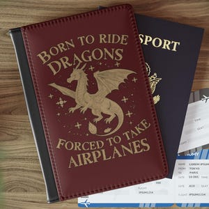 Peut inclure: Un porte-passeport bordeaux avec le texte "BORN TO RIDE DRAGONS FORCED TO TAKE AIRPLANES" et une illustration de dragon doré. Un passeport bleu foncé et deux billets d'avion sont à côté. Une clé de voiture est en arrière-plan.