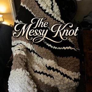 May include: A chunky knit blanket featuring thick, soft yarn in alternating horizontal stripes of cream, tan, and black. The text "The Messy Knot" is overlaid in white cursive script across the center of the textured, looped fabric surface.