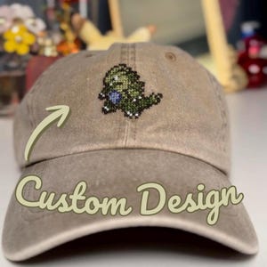 May include: A tan baseball cap with a cross-stitched dinosaur design. The cap features the words "Custom Design" in a cursive font. The dinosaur is green with a blue belly.