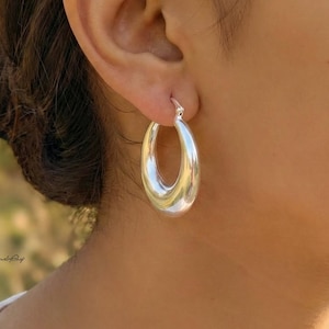 Puede incluir: Primer plano de un pendiente de aro plateado. El pendiente tiene una forma ovalada lisa con un acabado pulido. El pendiente se lleva en la oreja de una persona, con el cierre visible. El fondo está borroso, con un toque de verde.