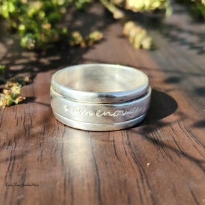 Peut inclure: Une bague argentée avec l'inscription gravée "I am enough". La bague a une bande rotative et est posée sur une surface en bois avec de la verdure en arrière-plan. C'est un bijou.