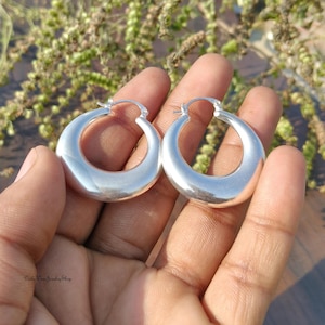 Puede incluir: Un par de pendientes de aro de plata pulida. Los pendientes tienen forma redondeada de media luna y una superficie lisa y reflectante. Los pendientes se sostienen en la mano de una persona sobre un fondo borroso de vegetaci&oacute;n.