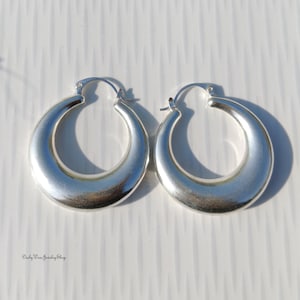 Puede incluir: Un par de pendientes de aro plateados. Los pendientes tienen forma redondeada de media luna con un acabado liso y pulido. Los pendientes se muestran sobre una superficie blanca.