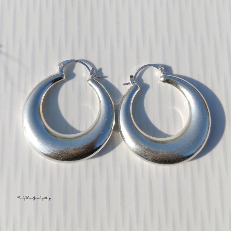 Puede incluir: Un par de pendientes de aro plateados. Los pendientes tienen forma redondeada de media luna con un acabado liso y pulido. Los pendientes se muestran sobre una superficie blanca.
