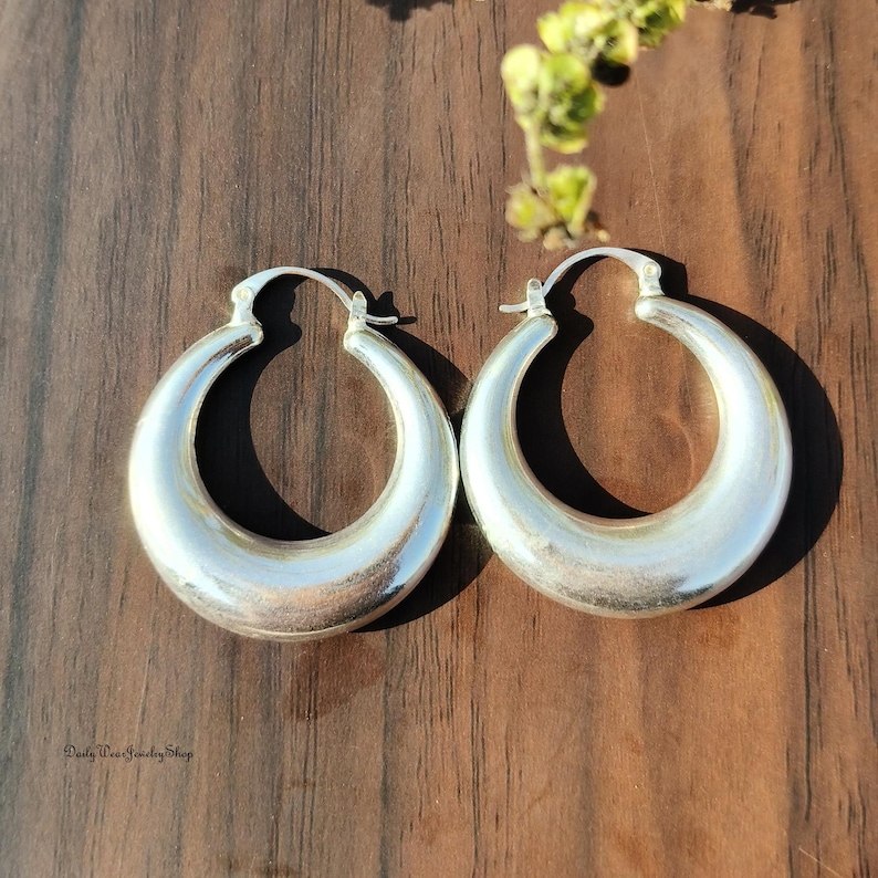 Puede incluir: Un par de pendientes de aro plateados con un acabado pulido. Los pendientes son gruesos y redondeados, con un dise&ntilde;o cl&aacute;sico. Los pendientes se exhiben sobre una superficie de madera oscura, con una ramita de vegetaci&oacute;n en el fondo. Los pendientes son de Daily Pea Jewelry Shop.