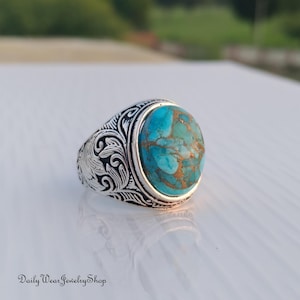May include: A silver ring with an oval turquoise stone. The stone has a mottled pattern of blue and brown. The ring band features an ornate, engraved design. The text "Daily Wear Jewelry Shop" is at the bottom of the image.