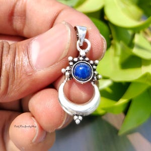 May include: A silver pendant with a deep blue gemstone center. The pendant features a crescent moon design with a circular gemstone setting and decorative accents. The Daily Wear Jewelry Shop logo is visible at the bottom.
