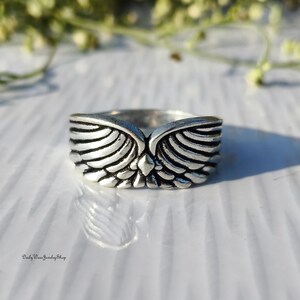 May include: A silver-colored ring with a detailed wing design. The ring features a central diamond-shaped accent and the text "Daily Wear Jewelry Shop" at the bottom. The ring is set against a white surface.
