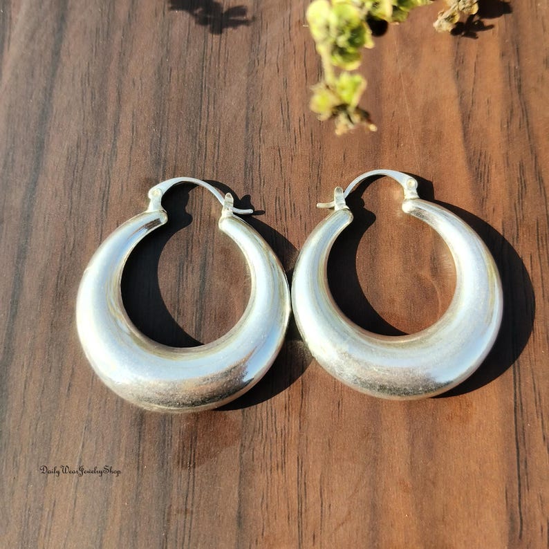 Puede incluir: Un par de pendientes de aro plateados. Los pendientes tienen un dise&ntilde;o grueso y redondeado con un acabado pulido. Se presentan sobre una superficie de madera oscura. El logotipo de Daily Wear Jewelry Shop es visible en la esquina inferior izquierda.