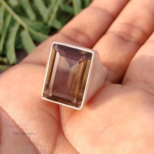 Könnte beinhalten: Ein rechteckiger Ring mit Rauchquarz-Edelstein und einem silbernen Band. Der Edelstein hat eine transparente, hellbraune Farbe und ist in einem schlichten, modernen Design gefasst. Der Ring wird auf einer Hand gezeigt.