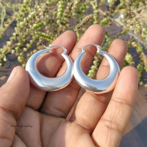 Puede incluir: Un par de pendientes de aro de plata pulida. Los pendientes tienen forma redondeada de media luna y una superficie lisa y reflectante. Los pendientes se sostienen en la mano de una persona sobre un fondo borroso de vegetaci&oacute;n.