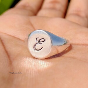 May include: A silver signet ring with a polished finish. The ring features a circular face with the letter "E" engraved in a cursive font. The ring is shown on a hand.