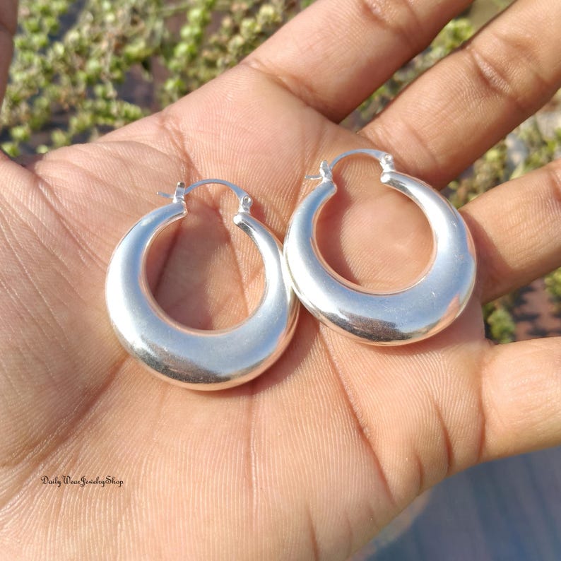 Puede incluir: Un par de pendientes de aro plateados. Los pendientes tienen una forma ovalada redondeada con una superficie lisa y pulida. Se sostienen en una mano. El texto "Daily Wear Jewelry Shop" es visible en la parte inferior izquierda.