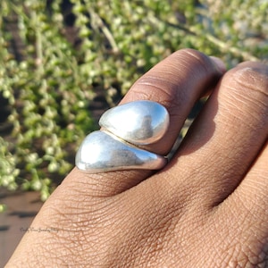May include: A silver ring with a distinctive, abstract design. The ring has two rounded, smooth shapes that overlap, creating a modern look. The ring is worn on a finger, showcasing its polished surface.