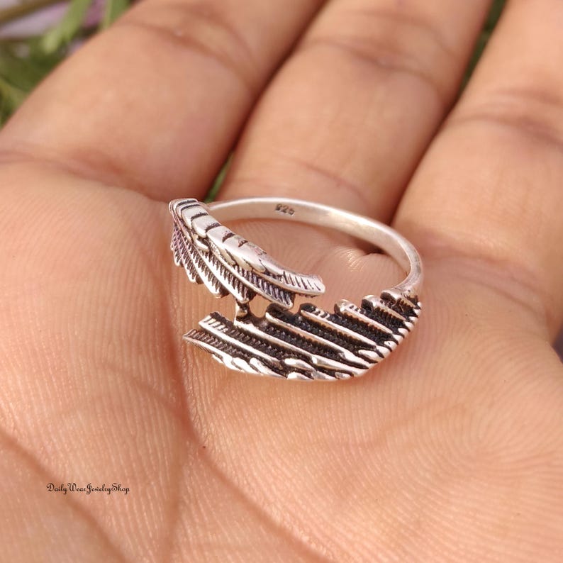 May include: A silver-colored ring with a feather design. The ring features a detailed feather motif, with intricate lines and textures. The ring is set against a warm, neutral background.