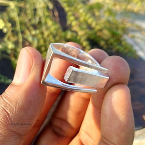 May include: A silver-colored ring with a geometric design. The ring features a rectangular bar that is connected to a square frame. The ring is held in a hand against a blurred green background.