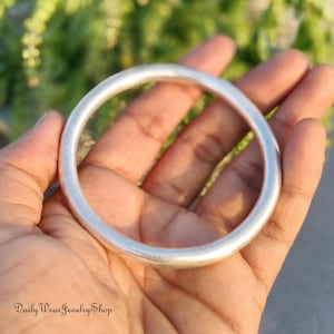 Puede incluir: Una pulsera de brazalete de color plateado con un diseño liso y redondeado. La joya se sostiene en una mano, con un fondo verde borroso. El texto "Daily Wear Jewelry Shop" es visible en la parte inferior izquierda.