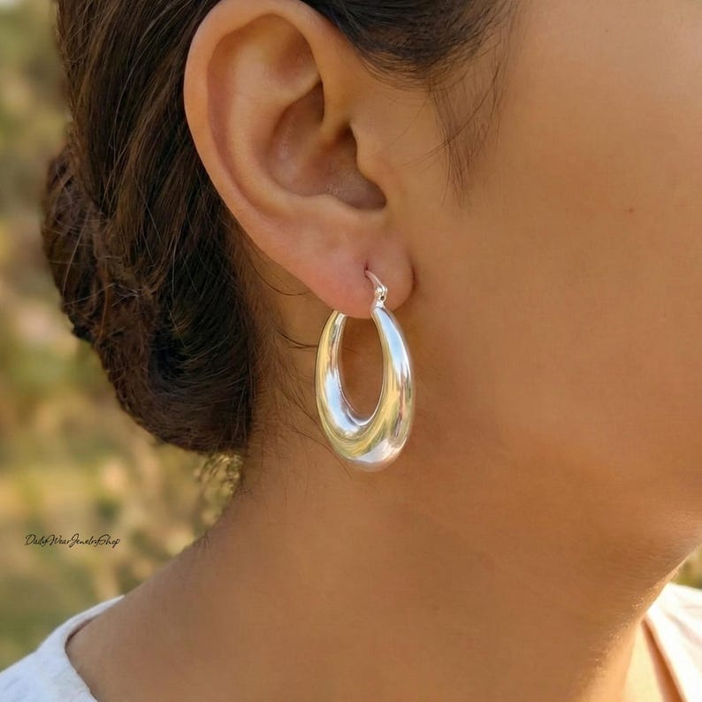 Puede incluir: Primer plano de un pendiente de aro plateado. El pendiente tiene una forma ovalada lisa con un acabado pulido. El pendiente se lleva en la oreja de una persona, con el cierre visible. El fondo est&aacute; borroso, con un toque de verde.