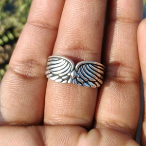 May include: A silver-colored ring with a detailed wing design. The ring features two wings that meet in the center, creating a unique and symbolic piece of jewelry. The ring is worn on a finger, showcasing its intricate details.