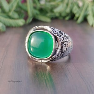 May include: A silver ring with a large, square, bright green stone. The ring has an ornate, engraved design around the stone and on the band. The ring is displayed on a reflective surface.
