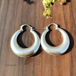 Puede incluir: Un par de pendientes de aro plateados. Los pendientes tienen forma de media luna redondeada con un acabado pulido. Los pendientes se muestran sobre una superficie de madera oscura. El texto "Daily Wear Jewelry Shop" es visible en la esquina inferior izquierda.