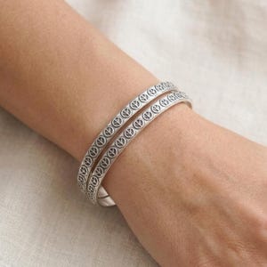 May include: Two silver-toned metal bangle bracelets featuring a repeating embossed peace sign pattern. The bracelets have a smooth, polished finish and a thin, rounded profile, worn stacked together on the wrist to create a layered appearance.