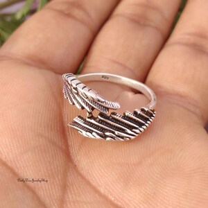 May include: A silver-colored ring with a feather design. The ring features a detailed feather motif, with intricate lines and textures. The ring is set against a neutral background, highlighting the ring's design. The ring is a piece of jewelry.