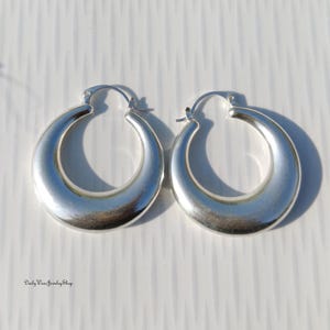 Puede incluir: Un par de pendientes de aro plateados. Los pendientes tienen forma redondeada de media luna con una superficie lisa y pulida. Los pendientes se muestran sobre una superficie blanca.