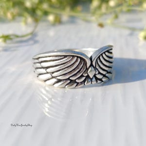 May include: A silver-toned ring with a detailed wing design. The ring features a textured surface with individual feather details, creating a three-dimensional effect. The wings are spread out, giving the ring a unique and elegant look.