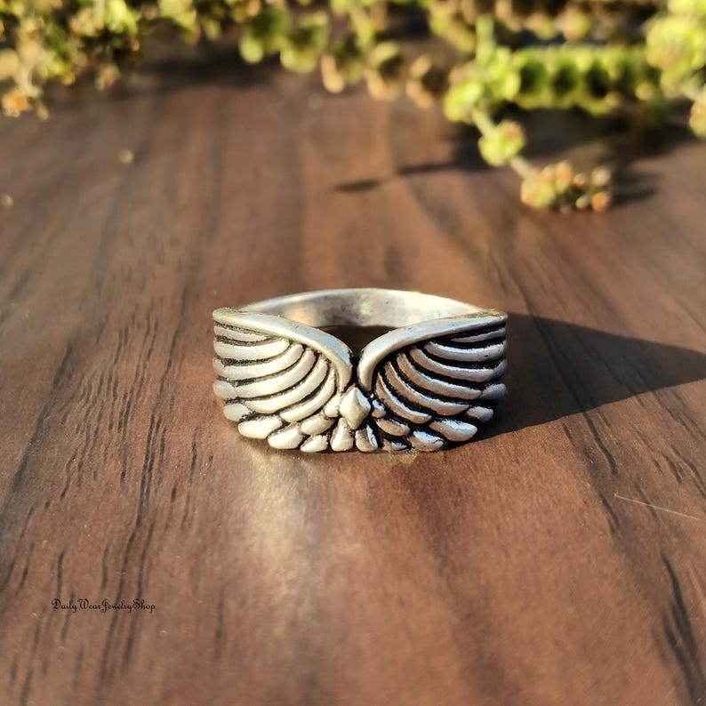 May include: A silver-toned ring with a wing design. The ring features detailed feather textures, with the wings extending from the center. The ring is set against a wooden surface, with a blurred background of greenery. The ring is a piece of jewelry.