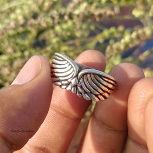 May include: A silver-colored ring with a detailed wing design. The ring features two wings extending outwards, with each feather meticulously crafted. The ring is held between fingers, showcasing the intricate details of the jewelry.