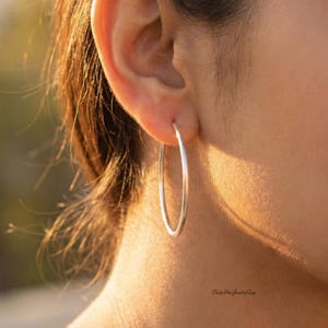 May include: A close-up of a silver hoop earring worn in an ear. The earring is a simple, polished metal design. The image is well-lit, highlighting the smooth surface of the jewelry. The background is blurred, focusing attention on the earring.