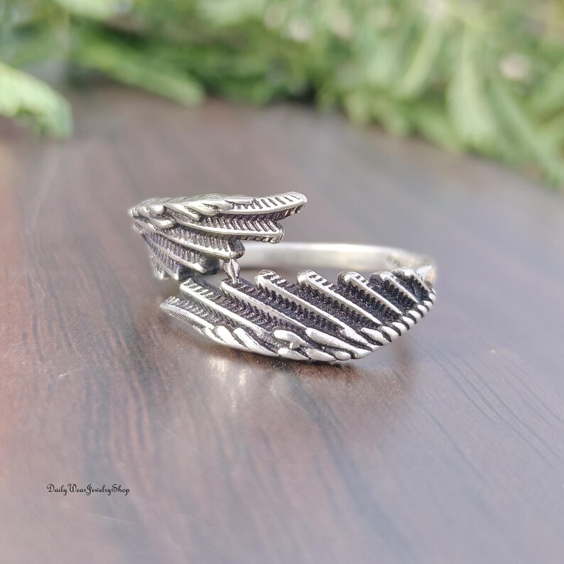 May include: A silver-colored ring with a detailed feather design. The ring wraps around the finger with the feather design on top. The ring is on a wooden surface with green foliage in the background. The ring is from Daily Wear Jewelry Shop.