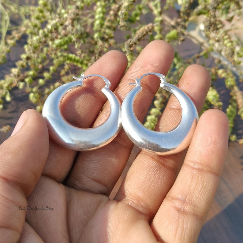 Puede incluir: Un par de pendientes de aro de plata pulida. Los pendientes tienen forma redondeada de media luna y una superficie lisa y reflectante. Los pendientes se sostienen en la mano de una persona sobre un fondo borroso de vegetaci&oacute;n.