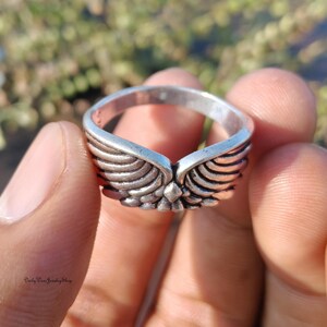 May include: A silver ring with a detailed wing design. The ring features a central point where the wings meet, and the wings are intricately carved with feather details. The ring is held in a hand, with a blurred background.