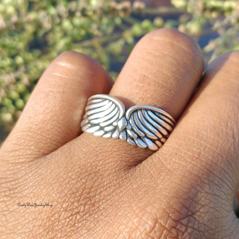 May include: A silver-toned ring featuring a detailed wing design. The ring's wings are spread out, with each feather meticulously crafted. The ring is worn on a finger, showcasing the intricate metalwork and design. The ring is from Daily Wear Jewelry Shop.