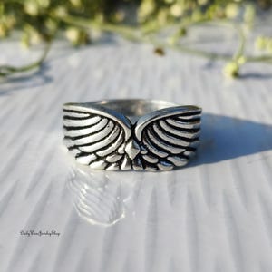 May include: A silver-colored ring with a detailed wing design. The ring features a central diamond-shaped accent and the text "Daily Wear Jewelry Shop" at the bottom. The wings are intricately carved, giving the ring a textured appearance.