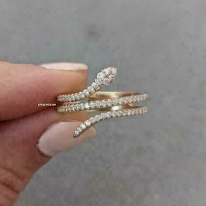 Può includere: Un anello a forma di serpente in oro rosa con un design avvolgente a tre fasce incastonate con piccole pietre chiare. L'anello riporta il testo KIVIJEWELSIN sul lato. Il metallo ha una calda tonalità rosata e una finitura lucida.