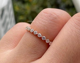May include: A thin rose gold band ring featuring a row of small, round, clear faceted stones set in a shared prong setting. The metal has a warm, metallic pink hue, and the stones are arranged in a continuous line across the top of the band.