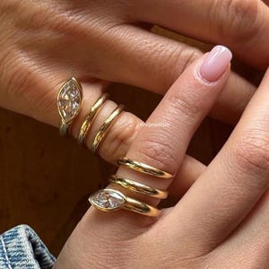 May include: A set of gold rings worn on fingers, featuring two rings with marquise-cut clear stones and four plain gold bands. The rings are arranged in a stacked style across the fingers, with the text Kivijewelsin visible in the centre.