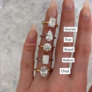 May include: Five rings with different diamond cuts arranged vertically. The labels read Emerald, Pear, Round, Radiant, and Oval. The rings feature various metal bands in gold and silver tones, each set with a single clear, faceted gemstone.