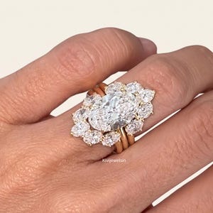 Puede incluir: Un anillo de oro con una gran piedra central de corte ovalado rodeada por un halo de piedras más pequeñas en forma de pera. La banda consta de tres hilos de oro paralelos que sostienen el engaste. El texto Kivijewelsin es visible en la banda.