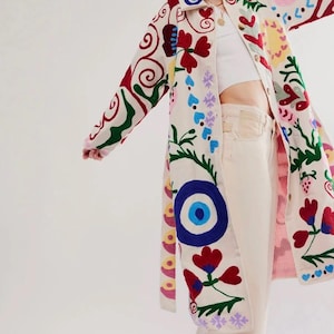 May include: A long, cream-coloured coat with vibrant, embroidered floral and geometric patterns in red, blue, green, and yellow. The coat features a large, eye-shaped design and is worn open, showcasing a white top and cream-coloured trousers.