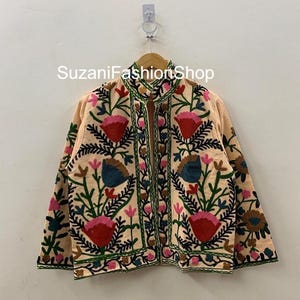 May include: A peach-colored jacket with intricate floral embroidery in red, pink, blue, and green. The jacket features a high collar and is adorned with detailed stitching, creating a vibrant and artistic design. The text "SuzaniFashionShop" is visible.