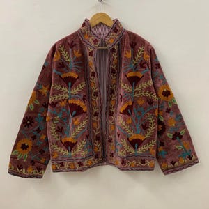 May include: Mauve velvet jacket with detailed floral embroidery in orange, yellow, and teal. The jacket has a high collar and long sleeves. The interior lining features a striped pattern.